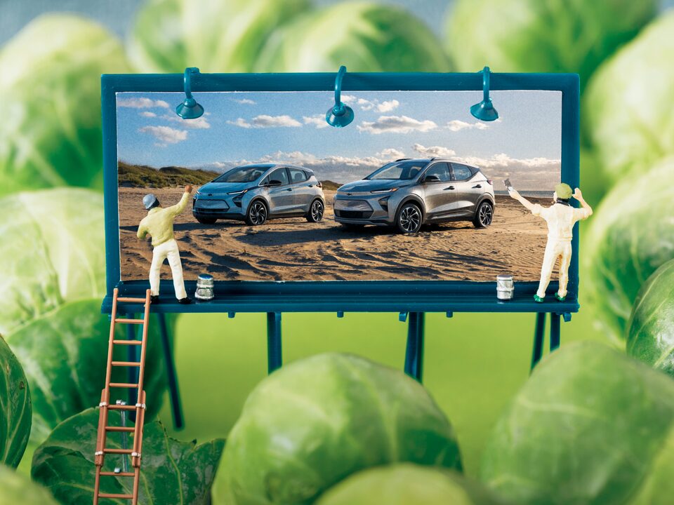 Una imagen tipo maqueta de una valla miniatura mostrando el Bolt EV y la Bolt EUV, con dos muñequitos pintándola, rodeada de coles de Bruselas para que parezcan árboles.