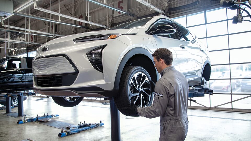 A service person works on a front wheel on a Chevy Bolt that is up on a hoist inside a service bay at a Chevy dealership.