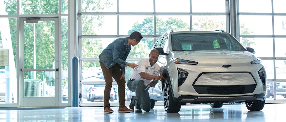 Dos personas conversan arrodilladas junto al neumático delantero de un Chevy Bolt en el taller de un concesionario de la marca.