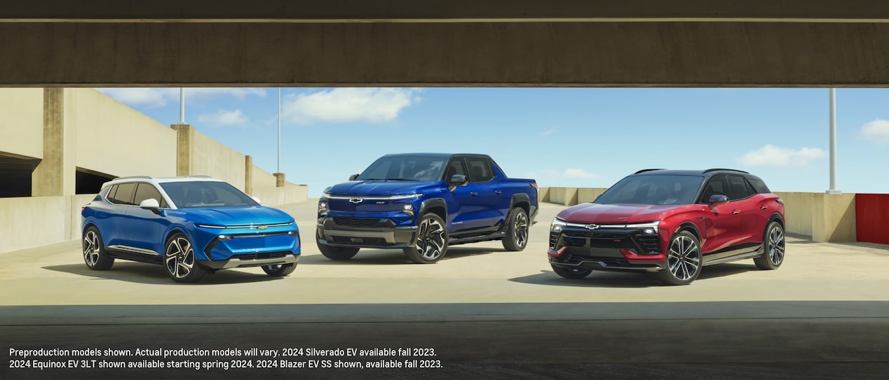 A blue Equinox EV, a blue Silverado EV, and a red Blazer EV are parked together on the open top level of a parking garage.