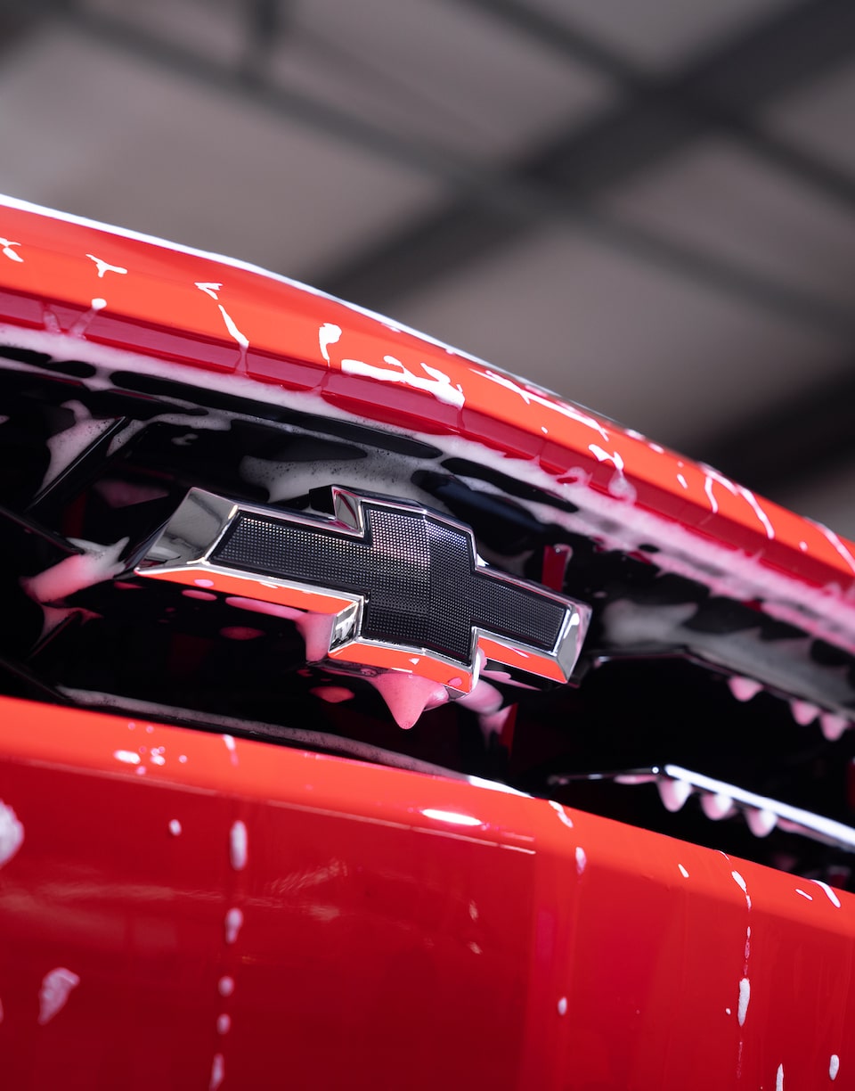 A close-up detail of the front grille of a red Chevy Camaro scattered with droplets of white foam.