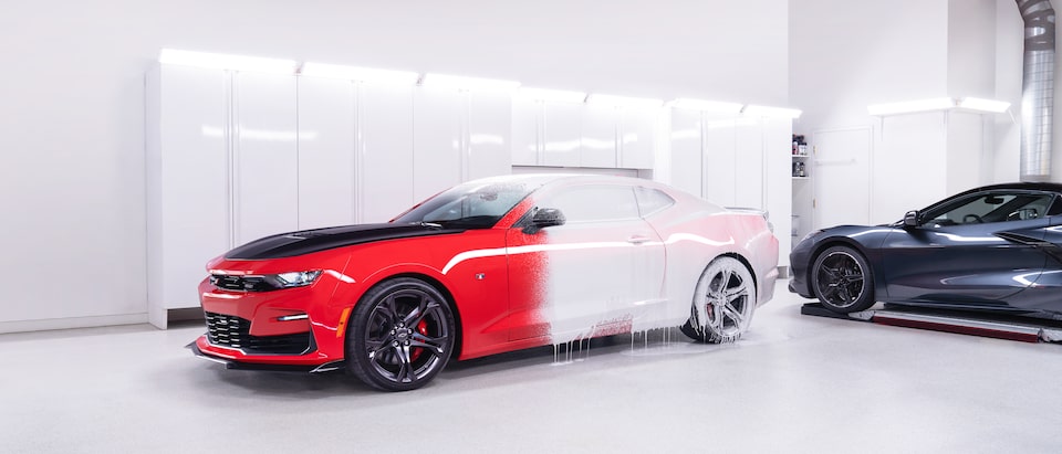 A bright red Chevy Camaro partly covered in a white foam sits in the bright white interior of the Esoteric Fine Auto Finishing shop next to a dark gray 2020 mid-engine Corvette.