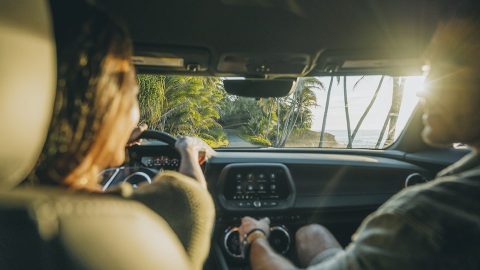 Vista delantera de una toma interior de una mujer y un hombre dentro de un Camaro SS 2020, con un paisaje de vegetación y el océano vistos a través del parabrisas.