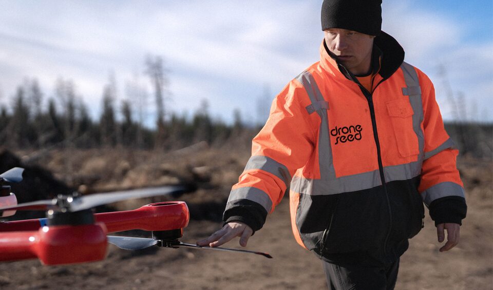 A person in an orange high-vis jacket stands in a treeless area with a large drone