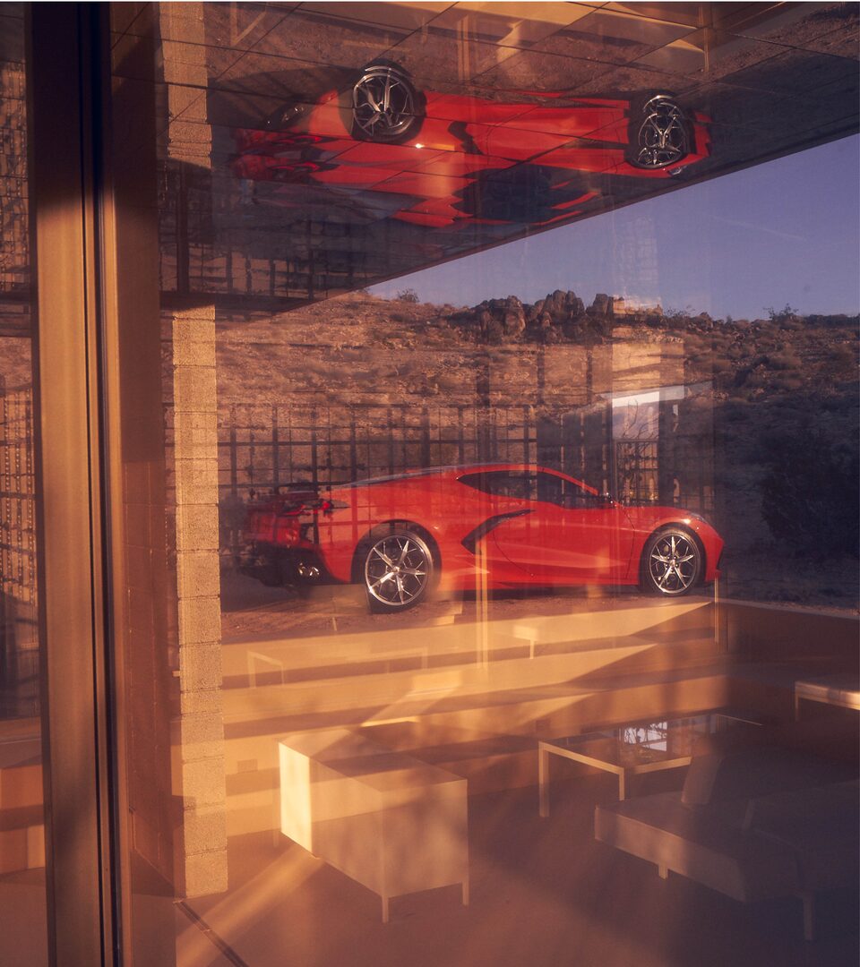 A Torch Red 2020 Corvette coupe sits in a desert landscape, seen through a window. A reflection of the car is in the glass.