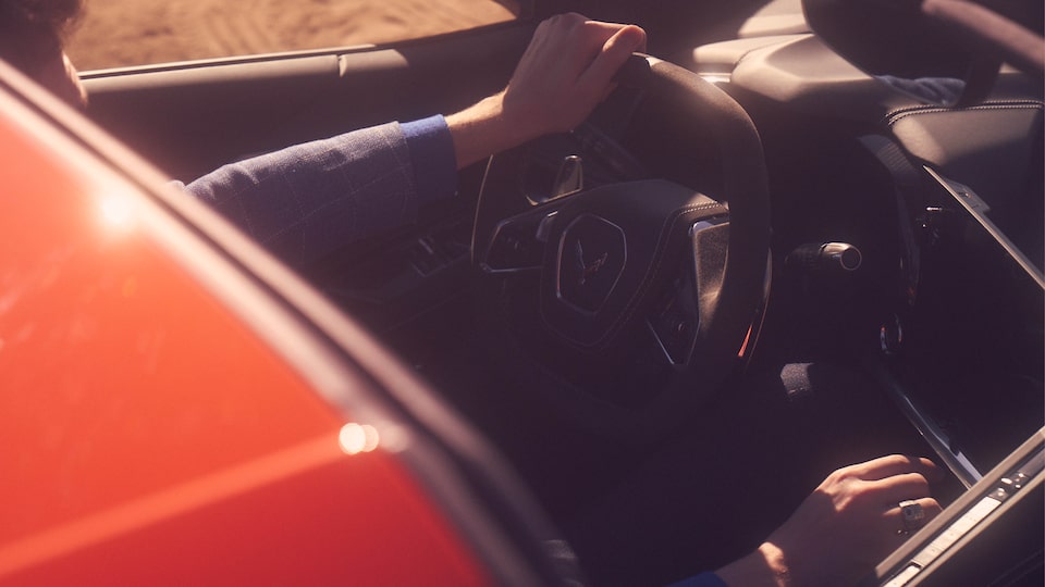 A view of the cockpit of the 2020 Corvette seen through the open roof. A person is in the driver’s seat, and the photo shows just one hand on the steering wheel and the other on the gear shift.