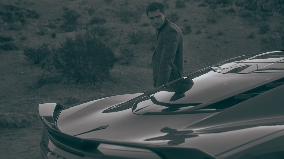 A man on the opposite side of a 2020 Corvette looks down at the rear of the car, seen from an angle slightly above the car to show off the rear windshield and back end.