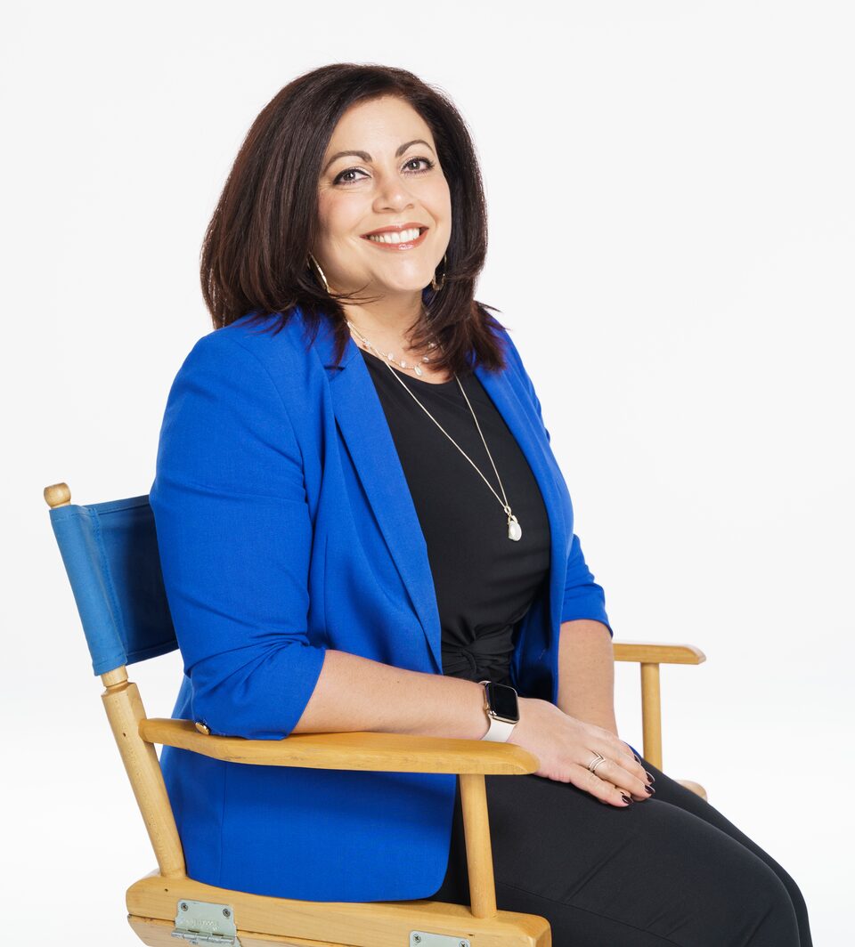 Jaclyn McQuaid, dressed in a navy shirt and lighter blue blazer sits on a blue director-style chair and smiles at the camera.