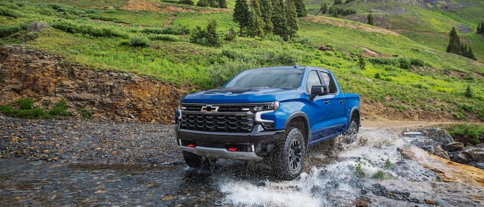 A blue Silverado ZR2 pickup truck crosses a gravel-covered stream bed. A dirt road and green hills and trees are in the background.