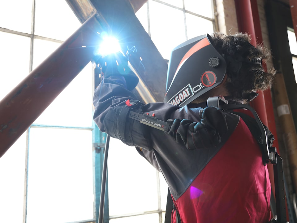 Una persona usando un exoesqueleto de alta tecnología y protector para el rostro suelda dos barras de hierro cruzadas frente a una ventana grande del tipo que se encuentran en depósitos.