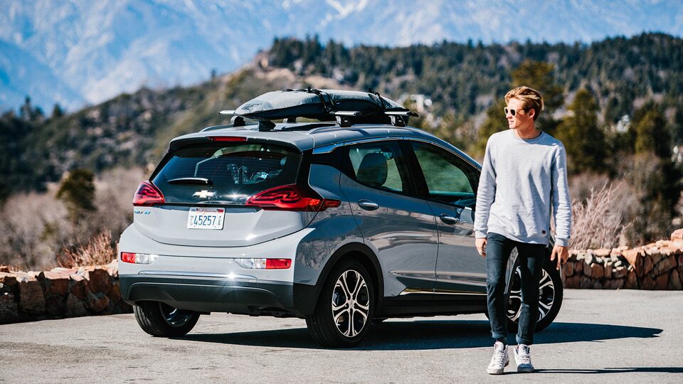 Kevin Schulz se aleja de un Bolt EV estacionado al costado de un camino de montaña con una tabla de surf y un snowboard sujetos a la parrilla portaequipajes en el techo.