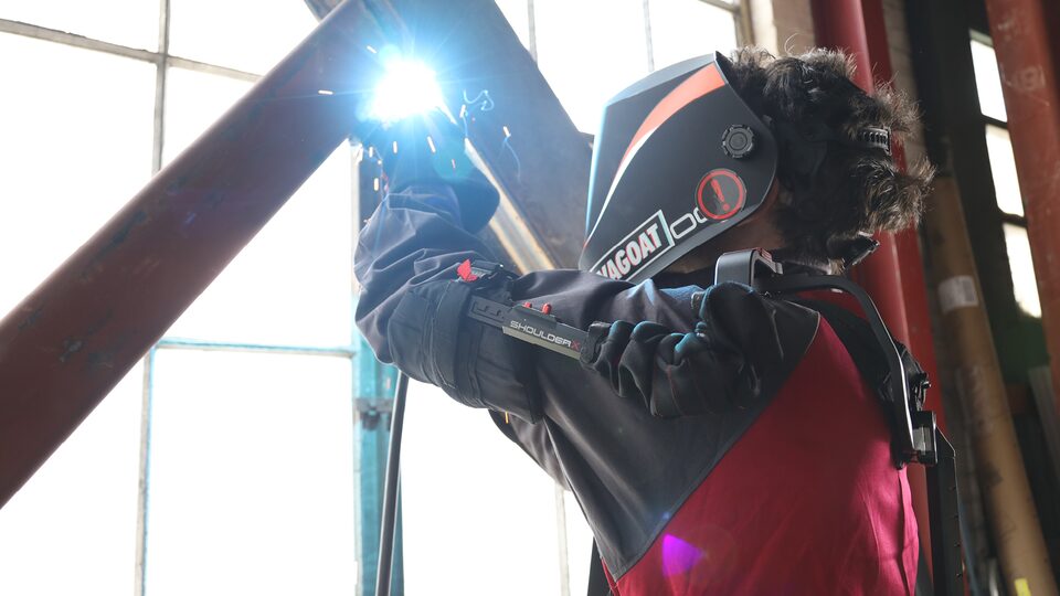 Una persona usando un exoesqueleto de alta tecnología y protector para el rostro suelda dos barras de hierro cruzadas frente a una ventana grande del tipo que se encuentran en depósitos.