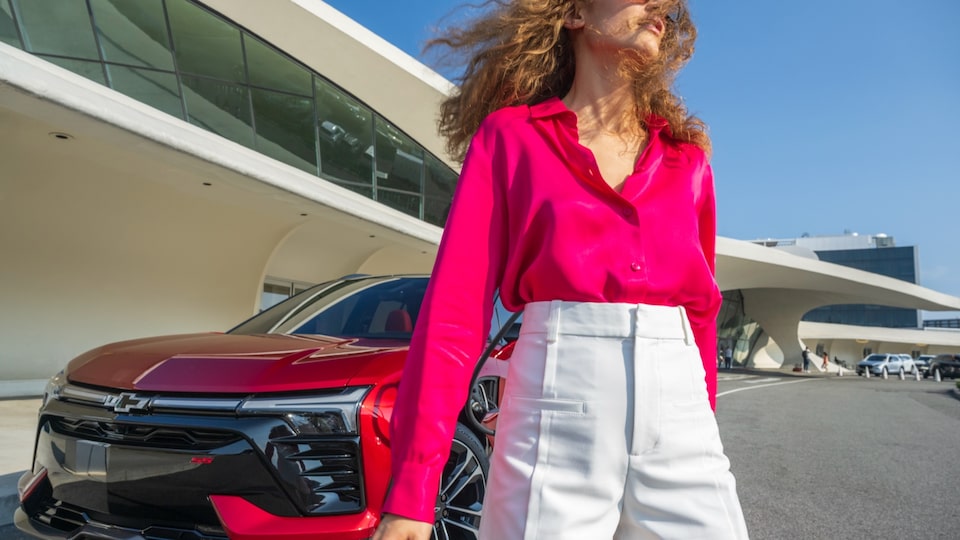 Una mujer con una blusa rosa brillante y pantalones cortos blancos se encuentra delante de una Blazer EV roja.