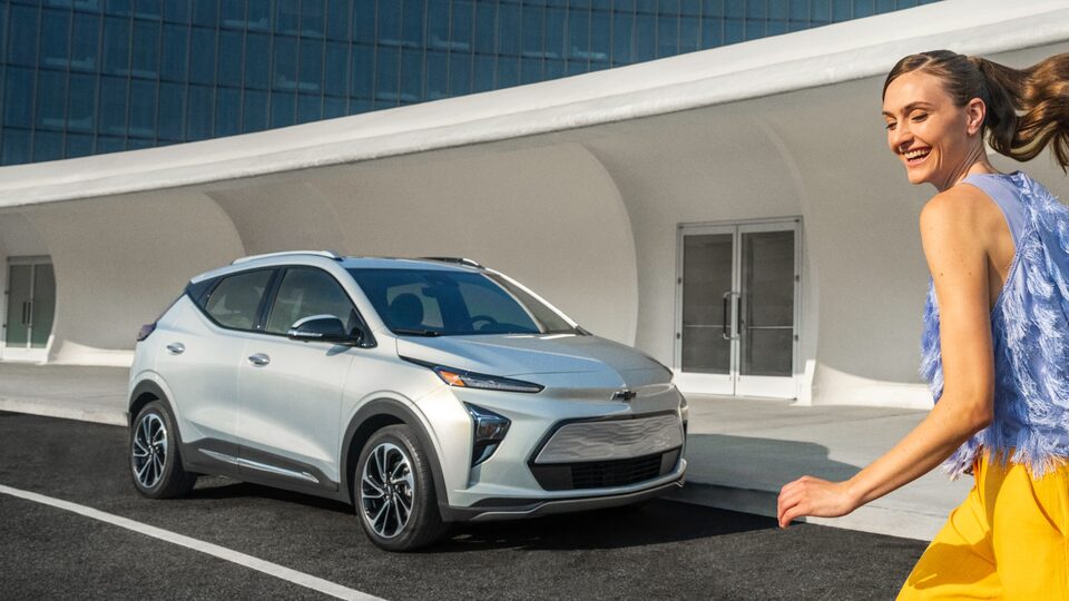 Una persona con pantalones cortos amarillos corre hacia una Bolt EUV plateada estacionada delante de un edificio de estilo contemporáneo.