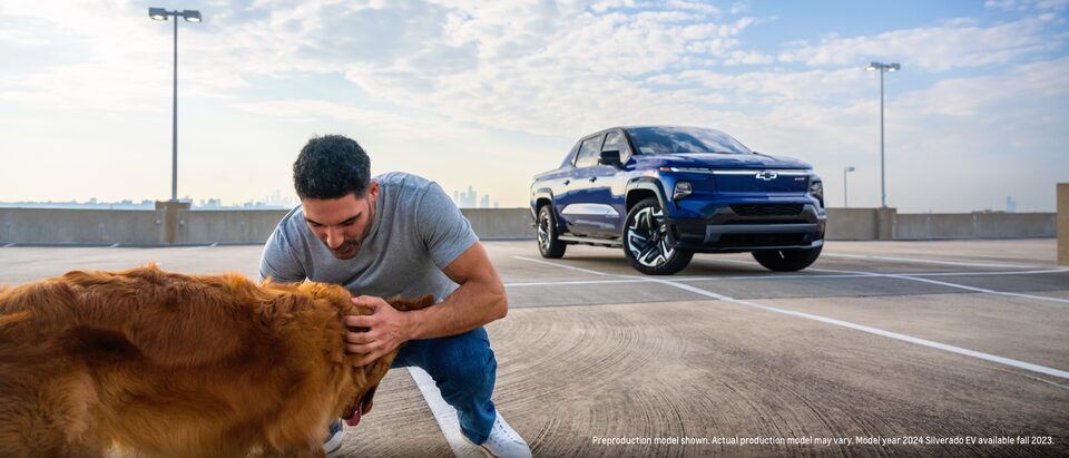A person kneels down to pet a Golden Retriever in a parking lot while a blue Silverado EV sits in the background.