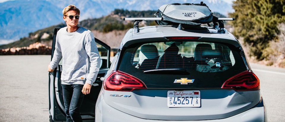 Kevin Schulz, wearing sunglasses, exits the driver’s door of a Bolt EV with a surfboard strapped to its roof rack.