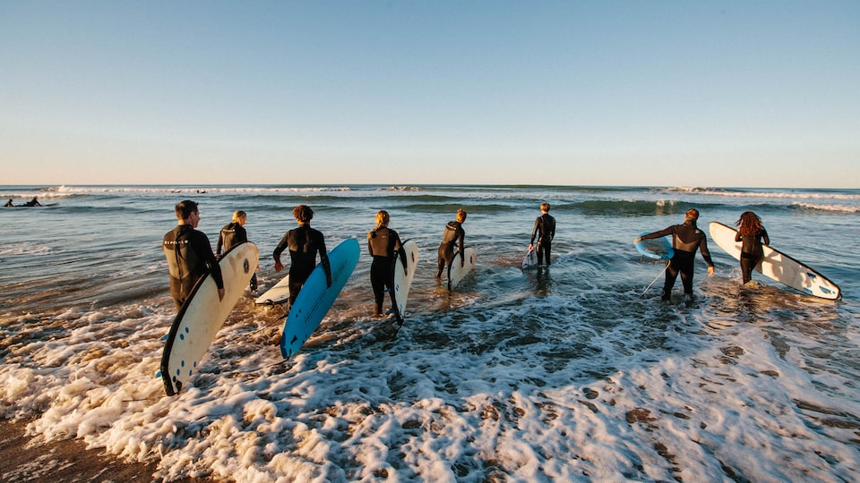 A group of people wearing wetsuits and carrying surfboards walk into the ocean.