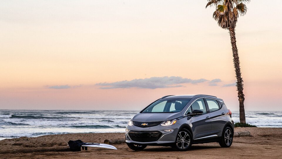 A Bolt EV sits on the beach with a single palm tree and the ocean in the background.