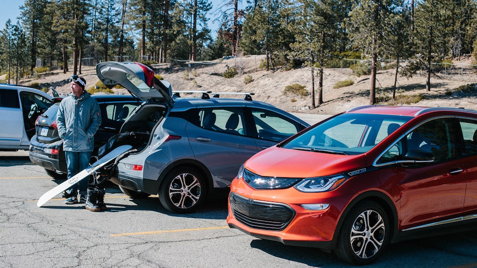 A line of Bolt EVs in a parking lot in the mountains. People are unloading a snowboard and other gear from the back of one of the Bolt EVs.