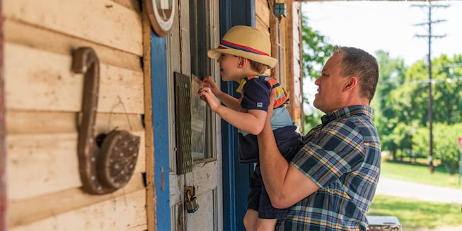 A man in a plaid shirt holds a young child up to look through a window in a building.