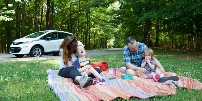 A man and woman and two young children sit on a colorful blanket on the grass having a picnic, with a white Bolt EV in the background.