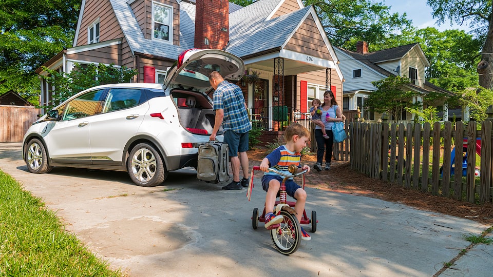 A man loads a suitcase into the open hatch of a white Bolt EV in a driveway in front of a brown house. A woman carrying a small child walks through a gate to the driveway while another child rides a tricycle in the driveway.
