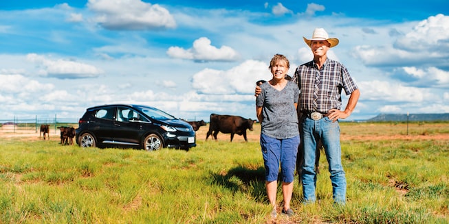 Un hombre y una mujer juntos en su rancho con un Bolt EV y vacas en el fondo