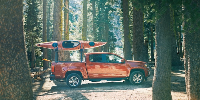 A Chevy Colorado with two kayaks on a gear rack drives through the forest.