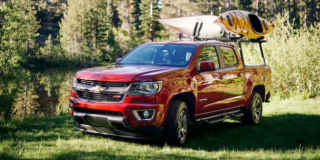 A Chevy Colorado with two kayaks on a gear rack parked next to the woods.