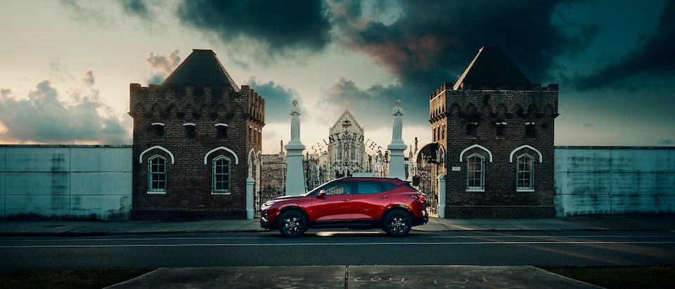 Una Chevy Blazer roja de perfil sobre una calle, frente a un portal de hierro forjado entre dos pilares grandes de ladrillo, con un cementerio de fondo y nubes oscuras en el cielo.