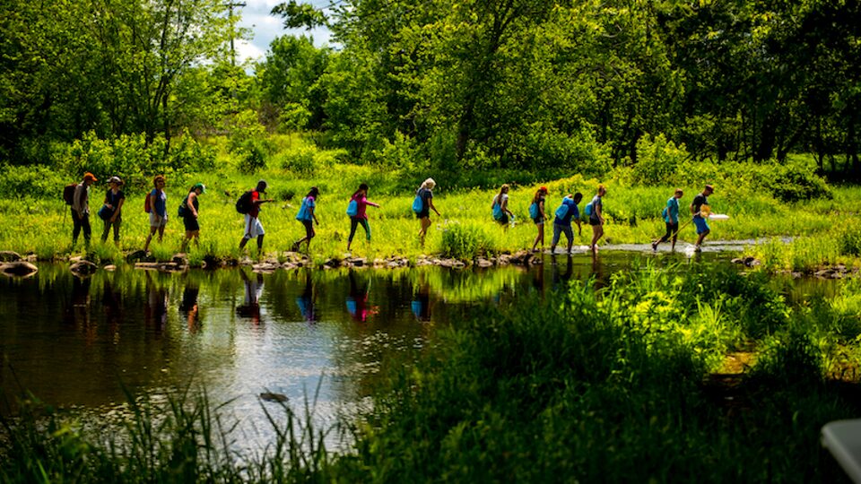 A group of students from the University of Maine Stormwater Management and Research Team walk in a single-file line next to a pond.