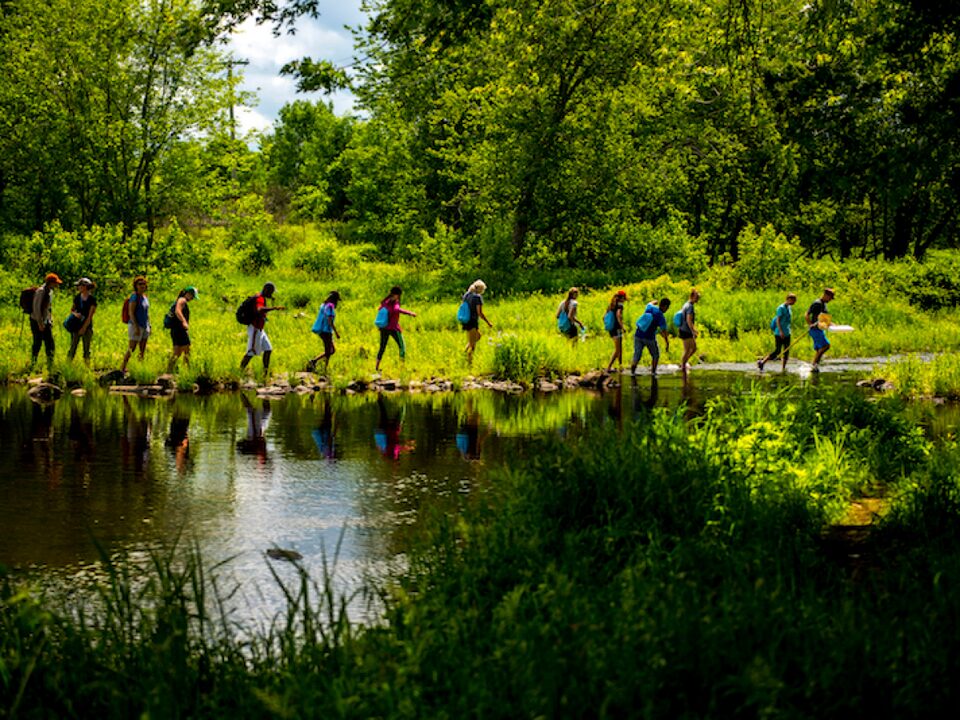 A group of students from the University of Maine Stormwater Management and Research Team walk in a single-file line next to a pond.