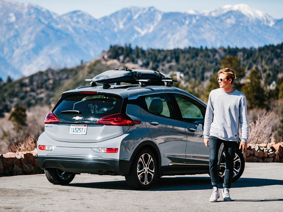 Kevin Schulz walks away from a Bolt EV parked on the side of a mountain road with a surfboard and snowboard strapped to the roof rack on top.