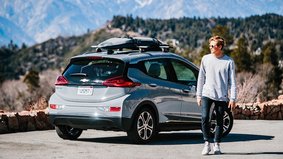 Kevin Schulz walks away from a Bolt EV parked on the side of a mountain road with a surfboard and snowboard strapped to the roof rack on top.