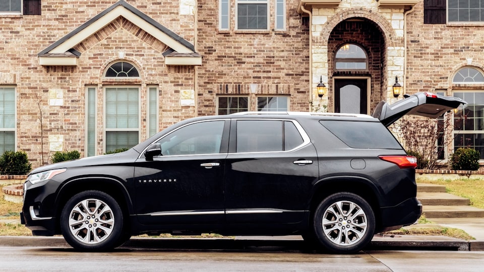 A black Chevy Traverse is parked curbside in front of a brick building with its back hatch open.