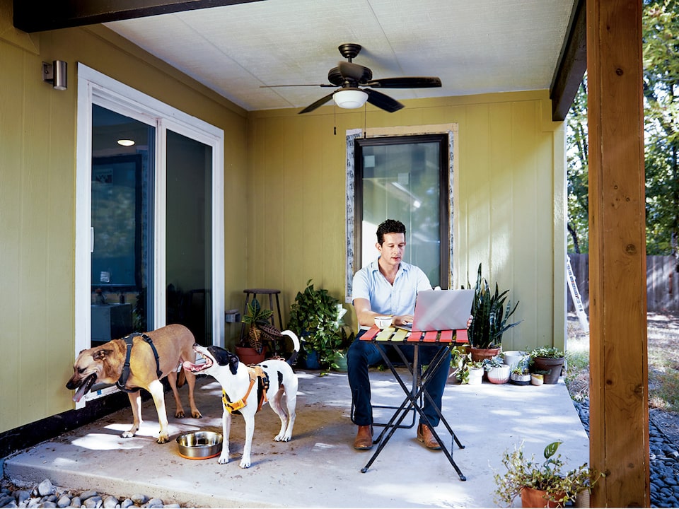 El arquitecto Jon Hagar usa una laptop sentado en una mesa que se encuentra en un patio cubierto y sus dos perros están sentados a su lado.