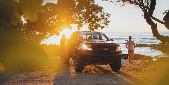 A red Chevrolet Colorado ZR2 is parked on the beach at sunset. One man is resting against the truck while the other is walking toward the water.