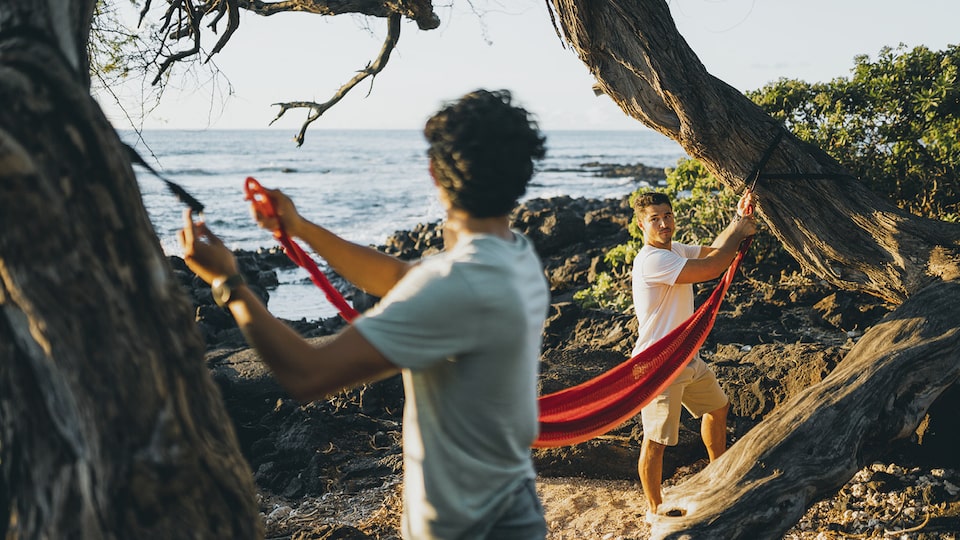 Two men attach a red hammock to opposite trees on the beach, with the water in the background.