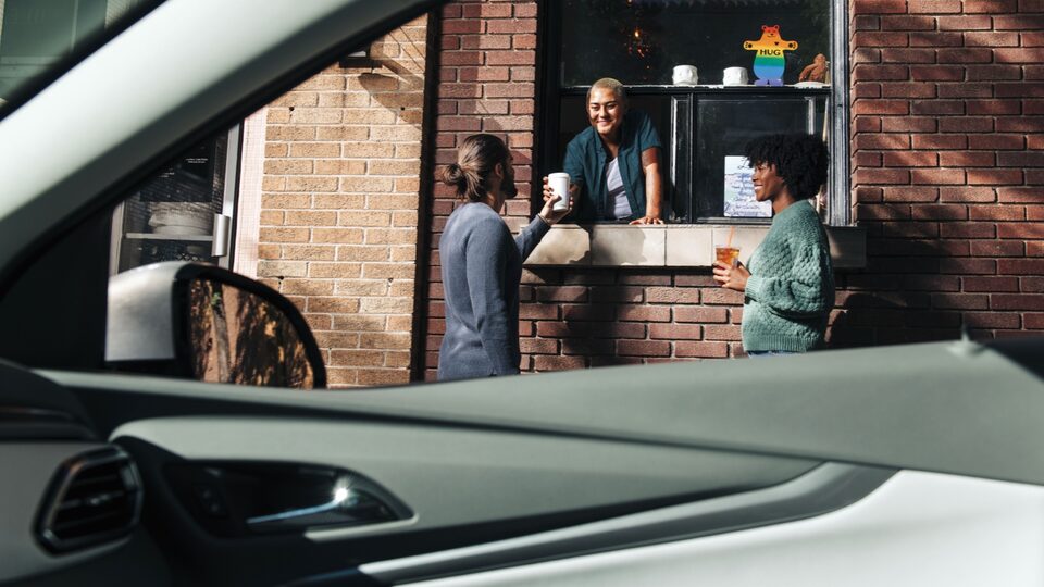 Two people stand at an outside service window of a coffeeshop, getting coffee and talking to the server inside.