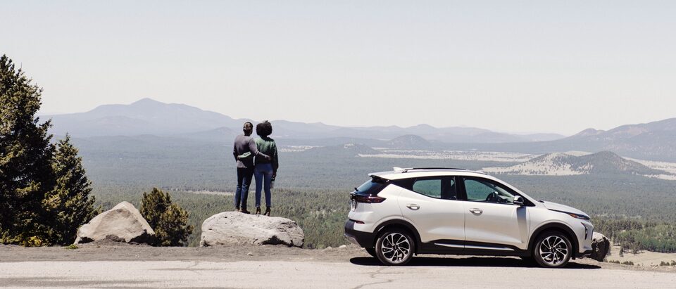 A white Chevy Bolt EUV sits in a pullout on a road in northern Arizona with a mountain landscape in the background. Two people stand on a large boulder next to the car.