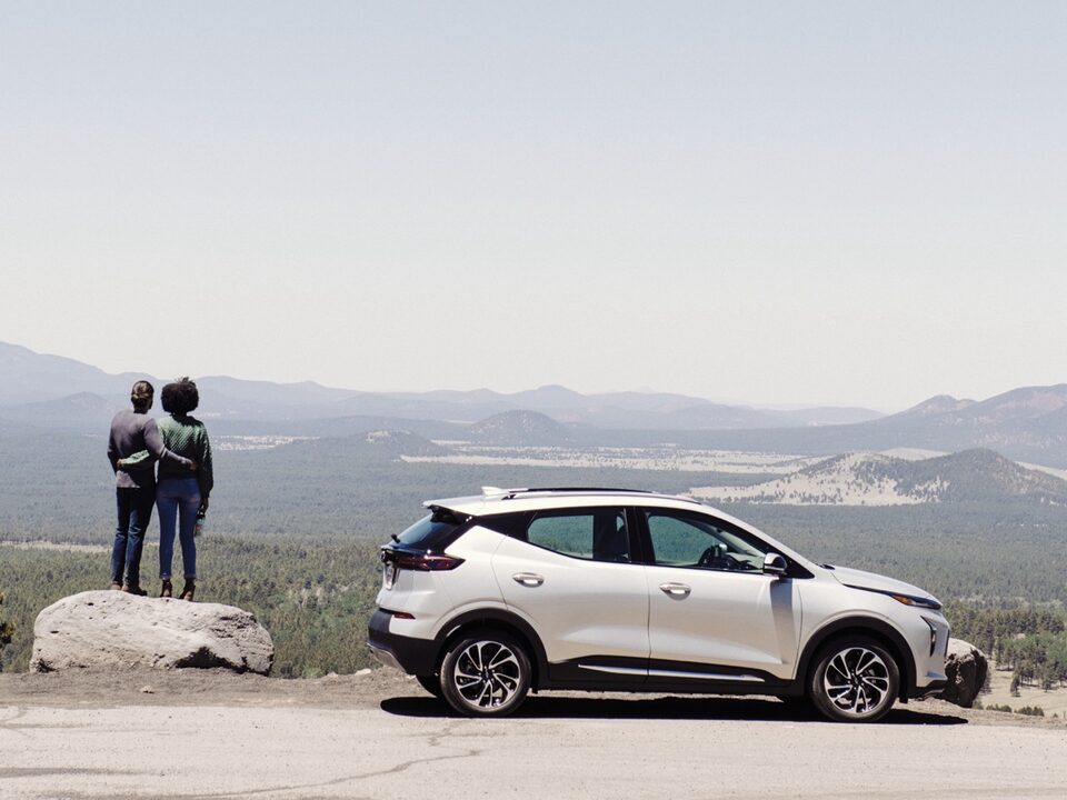 A white Chevy Bolt EUV sits in a pullout on a road in northern Arizona with a mountain landscape in the background. Two people stand on a large boulder next to the car.