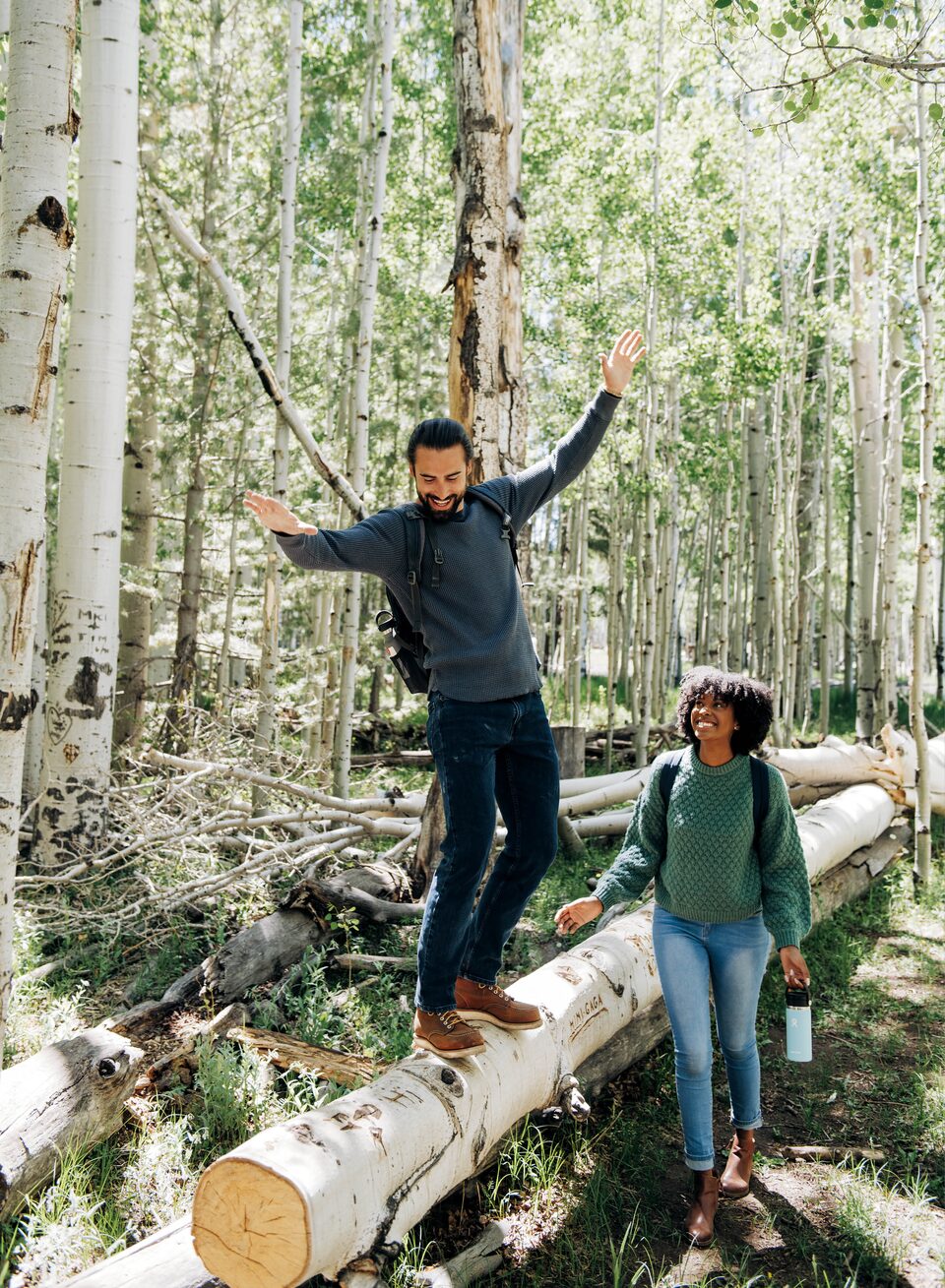 On a hiking trail in a forest, one person balances on a large log while another person watches.