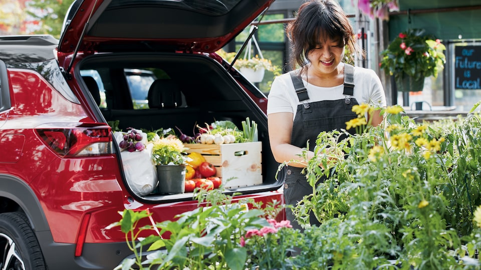 La chef Kim sonríe mientras observa un despliegue de flores amarillas en un mercado agrícola. Detrás de ella, una Chevy Trailblazer roja estacionada con el maletero trasero abierto con el área de carga llena de frutas, verduras y flores.