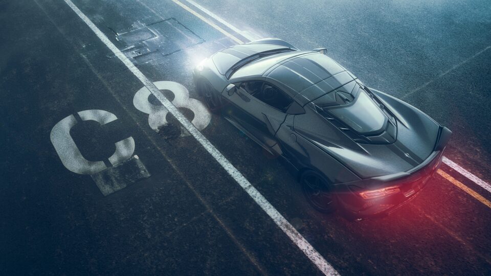 Un Corvette Stingray negro en miniatura visto desde arriba y con un brillo rojo proveniente de sus faros traseros, que parece estar moviéndose, con pavimento negro y marcas de carril de fondo, y con un esténcil de "C8" en blanco al lado del auto.