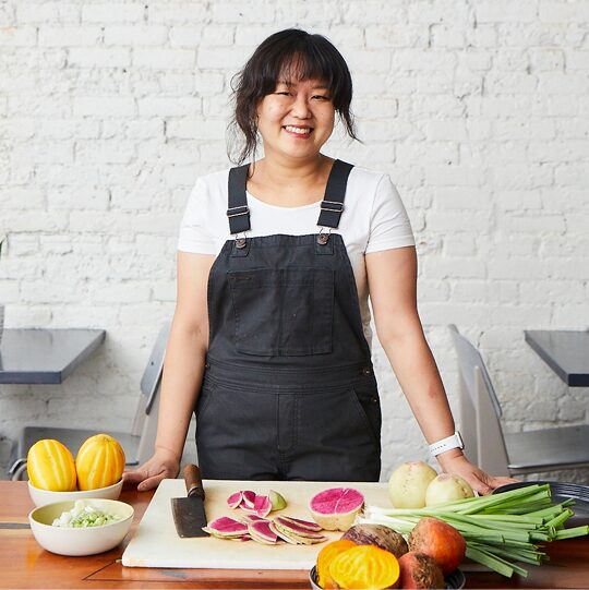 La chef Ji Hye Kim sonrié junto a la larga mesa de madera de su restaurante. En la mesa frente a ella, hay una tabla de cortar y una cuchilla con una gran variedad de verduras frescas.