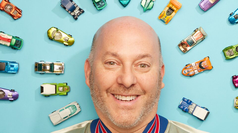 A head shot of Bruce Pascal in front of several Hot Wheels cars against a blue background.