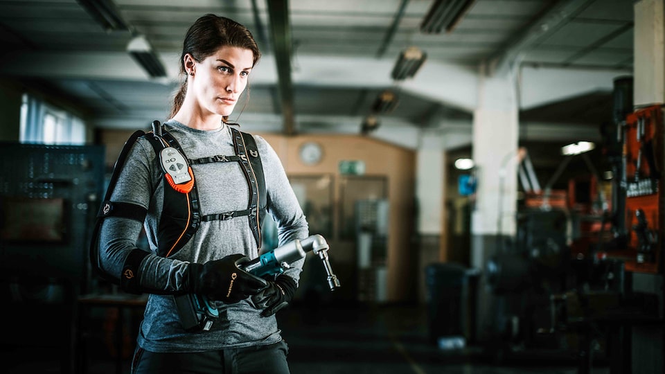 A woman wearing a high-tech exoskeleton suit on her upper body and high-tech gloves holds a tool in her right hand.