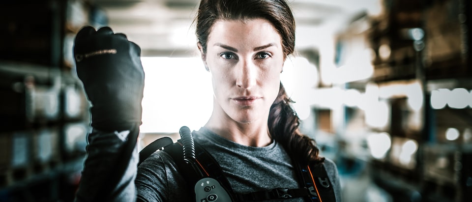A close-up shot of a woman wearing high-tech gear, showing her head and torso, as she looks directly at the camera and holds up her right hand to display a black glove.
