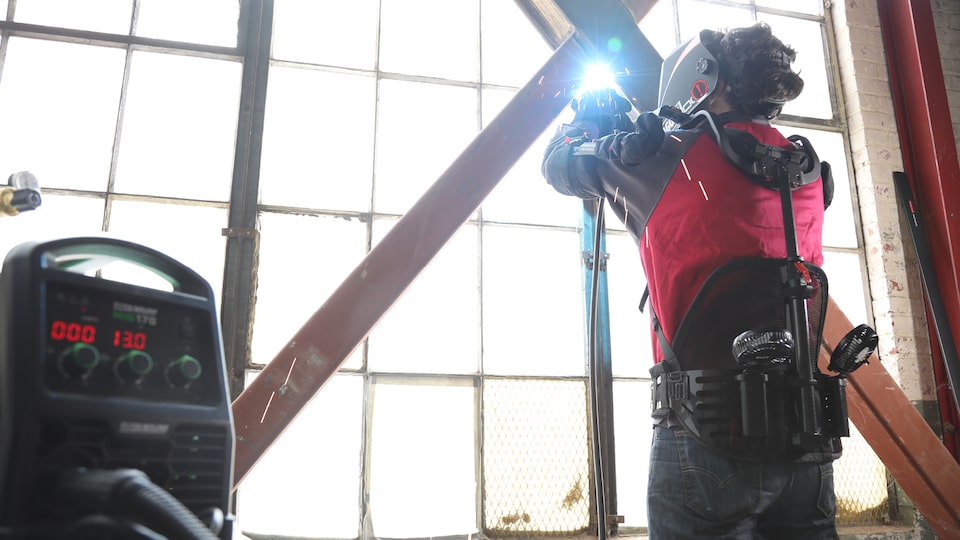 A person wearing a high-tech exoskeleton suit and a face shield welds two iron bars that cross in front of a large warehouse-type window.
