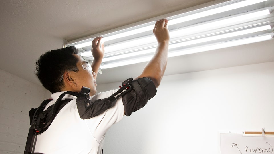 A man wearing a high-tech exoskeleton suit on his upper body installs fluorescent lights on a ceiling.